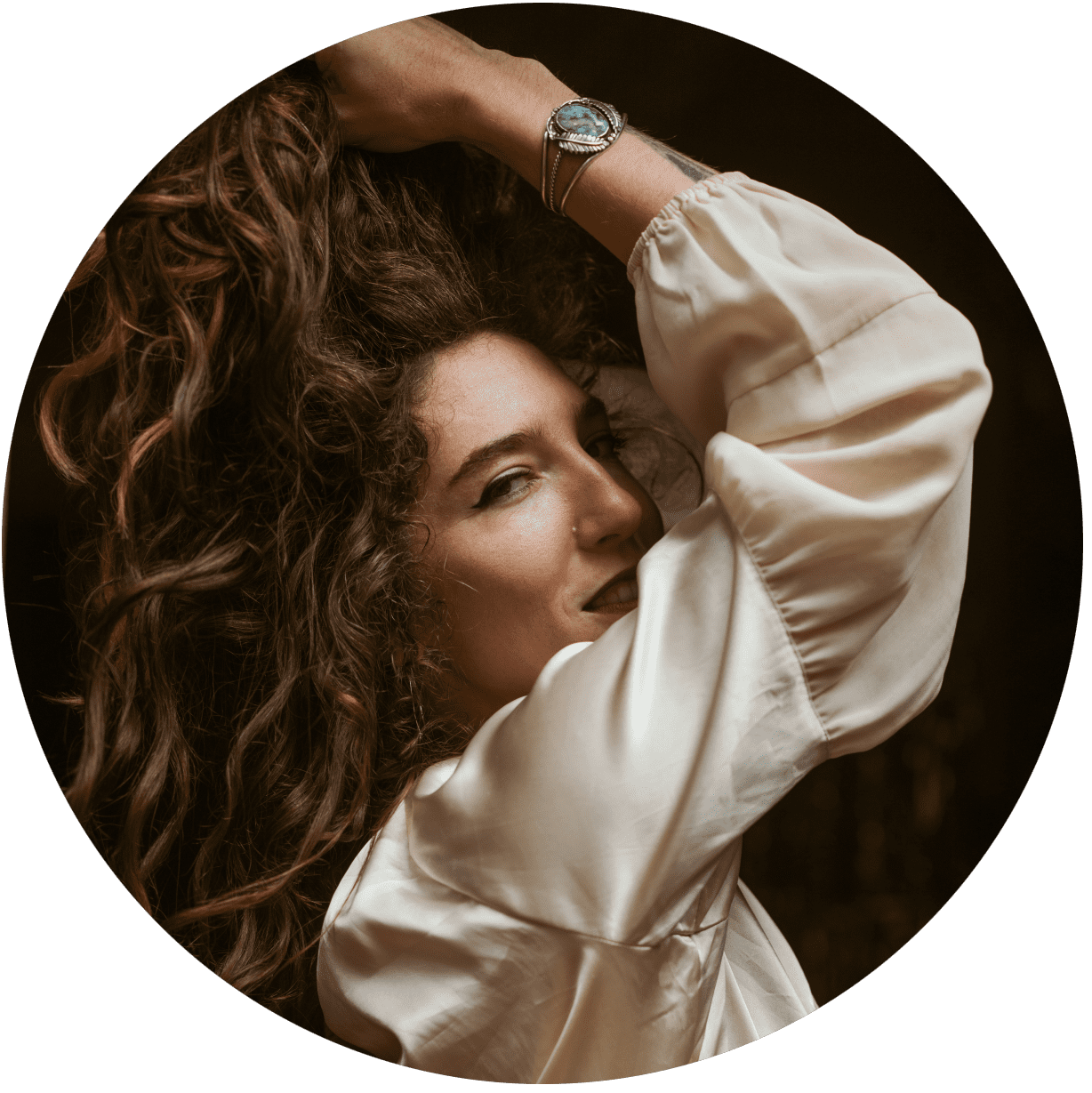 A woman with long curly hair wearing a light satin blouse poses with one arm raised, showing a watch on her wrist, and gazes confidently at the camera against a dark background.