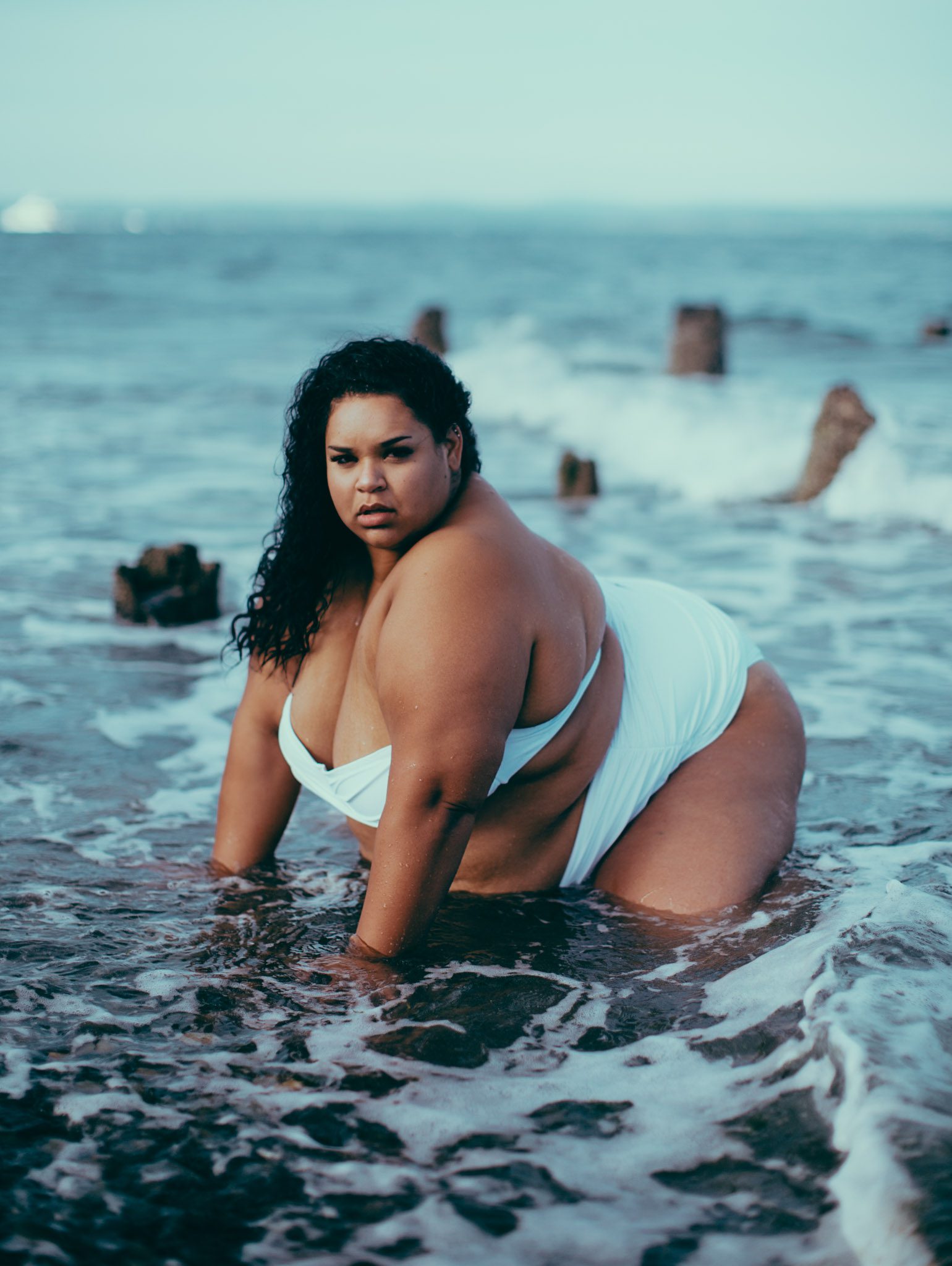 A woman in a white swimsuit poses confidently on her hands and knees in shallow ocean water, with waves and wooden posts in the background under a clear sky.